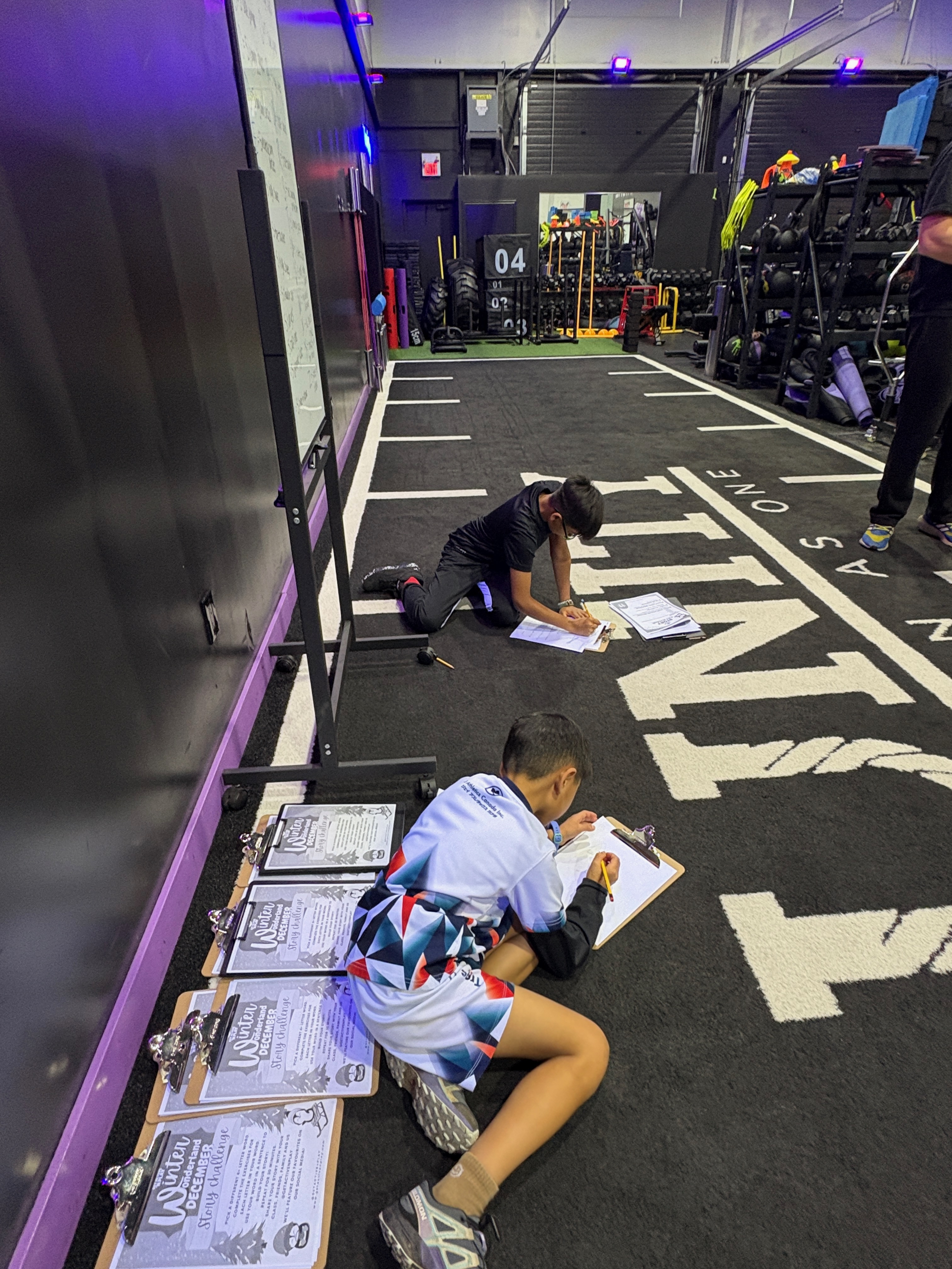 Two kids lying on the gym floor writing their stories on clipboards
